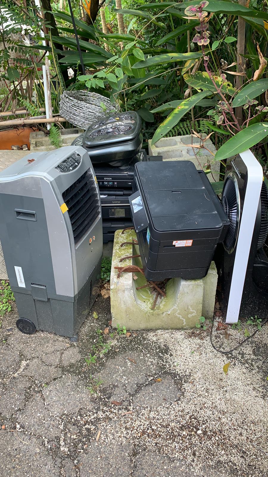 Pile of discarded electronics including an air cooler, printers, and fan stacked outdoors near greenery.